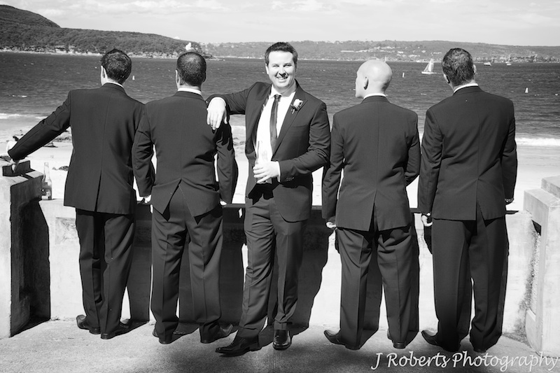 B&W of groom facing camera with groomsmen looking out to sea - wedding photography sydney